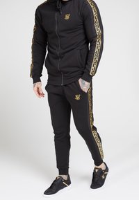 Felpa nera con zip e logo dorato, abbinata a pantaloni jogger neri con strisce laterali dorate. Sneakers nere con dettagli dorati.