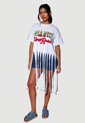 Frau mit dunklem Haar, die ein weißes T-Shirt mit Fransen trägt, auf dem "Wild West First Rodeo" steht, Denim-Shorts und blaue Sandalen mit offenem Zeh gegen einen einfarbigen Hintergrund.