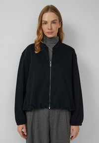 Czarna bomberka na zamek z elastycznymi mankietami i dołem, wykonana z miękkiego materiału, zestawiona z szarym topem z wysokim kołnierzem i eleganckimi spodniami.