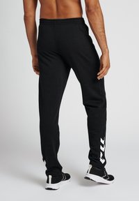 Zwarte joggingsbroek met elastische tailleband, taps toelopende benen en witte merklogo-accenten die verticaal langs de zijkant lopen. Draagt het met zwarte sneakers.