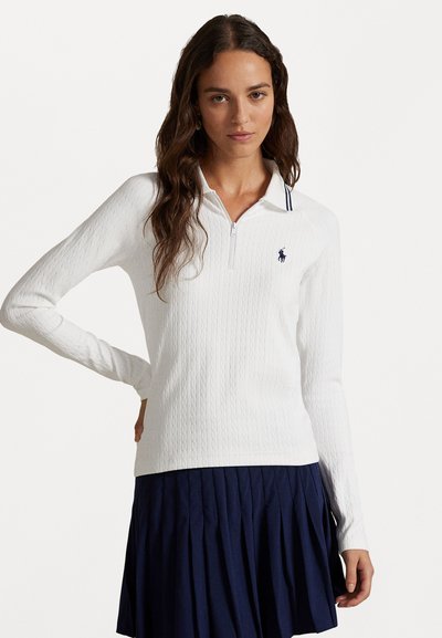 Weißes Langarm-Polo mit strukturiertem Kabelstrickmuster, Reißverschlusskragen und Akzent in Marineblau. Kombiniert mit einem marineblauen gefalteten Rock.