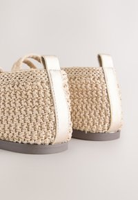 Gros plan de chaussures plates tissées beige avec des brides métalliques et des semelles grises sur fond clair.