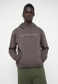 Hanorac maro cu buzunar frontal, glugă ajustabilă cu șnur și logo-ul "TOMMYJEANS" în alb. Modelul poartă pantaloni verzi.