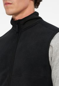 Άνδρας φοράει μαύρο fleece γιλέκο πάνω από ανοιχτό γκρι μακρυμάνικο πουκάμισο, με το γιλέκο φερμουδαρισμένο μέχρι το λαιμό.