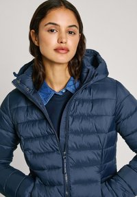 Mornarska puhovka z zaključenim dizajnom, kapuco in zadrgo. Nošena preko modre denim srajce in mornarske puloverja, ki prikazuje gladko teksturo.