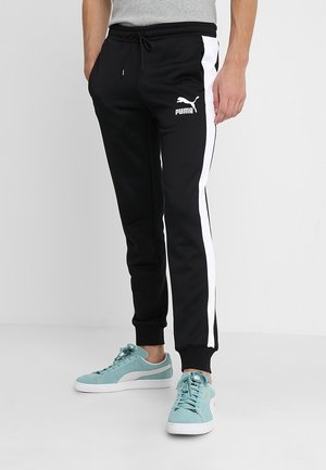 Mężczyzna w czarnych spodniach dresowych Puma z białymi paskami po bokach i turkusowymi butami Puma, stoi z rękami w kieszeniach.