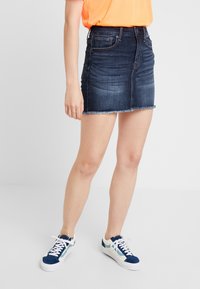 Osoba v tmavě modré denimové mini sukni s třepeným lemem, v kombinaci s jasně oranžovým topem a modro-bílými teniskami, stojící uvnitř.