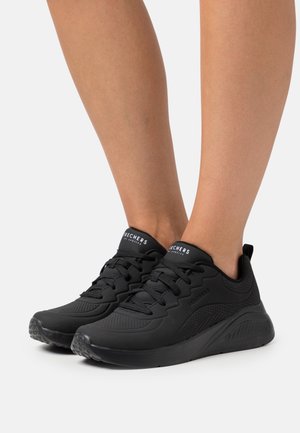 Voeten met zwarte Skechers veter- en sportschoenen met dikke zolen op een effen witte achtergrond.