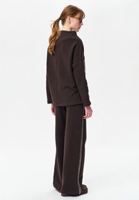 Bruine trui met een hoge kraag en ontspannen pasvorm, gecombineerd met bijpassende wijde broekspijpen met een decoratieve bies aan de zijkanten.