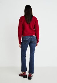 Suéter rojo texturizado de manga larga, combinado con jeans de campana oscuros y zapatos de tacón burdeos, visto desde la parte posterior.