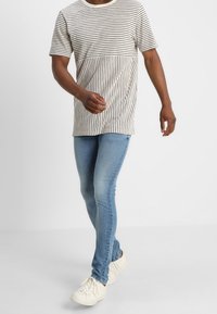 Camiseta de manga corta a rayas en beige y negro, combinada con jeans ajustados azules y zapatillas blancas. Conjunto sencillo y casual.
