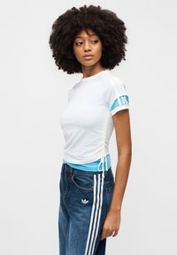 Λευκό αθλητικό τοπ με μπλε λεπτομέρειες, με σούρες στο πλάι και κοντά μανίκια. Συνδυασμένο με τζιν παντελόνι με μπλε ζώνη, τονισμένο με λευκές ρίγες.
