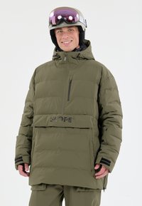 Grøn pufferjakke med horisontal quiltet design, frontlomme med lynlås, SLOPE-logo og justerbar hætte. Båret med hjelm og briller.