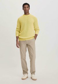 Gelb gestrickter Pullover mit geripptem Muster, kombiniert mit beigen, geraden Hosen und weißen Sneakers. Einfaches, lässiges Outfit.