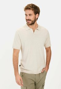Beżowa koszulka polo z krótkim rękawem, w desenie v-neck oraz z detalem brandingowym po bokach, zestawiona z oliwkowymi spodniami cargo wykonanymi z fakturowanej tkaniny.