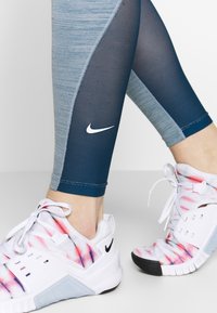 Modré legíny Nike s texturovaným tmavě modrým panelem a bílým logem, které jsou doplněny bílými teniskami s růžovým a modrým vzorem.