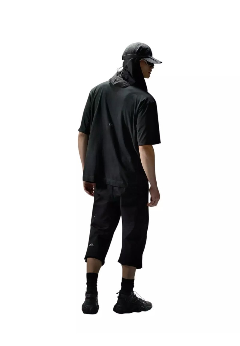 Homme portant un t-shirt noir oversize, un short mi-genoux, des chaussettes noires, des baskets, une casquette avec protection de nuque et des lunettes de soleil, debout, dos à la caméra.