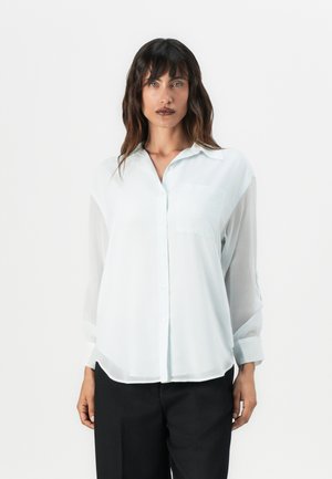 Blusa azzurro chiaro a maniche lunghe realizzata in tessuto trasparente con colletto, taschino anteriore e chiusura a bottoni. Indossata sopra pantaloni neri.