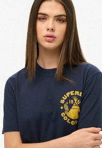 Camiseta azul marino de algodón, con cuello redondo, mangas cortas y un logotipo gráfico amarillo en el lado izquierdo del pecho.