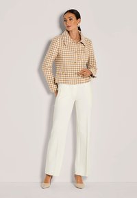 Vrouw in een beige houndstooth jas met knopen en witte wijde broek, poserend tegen een effen lichte achtergrond.