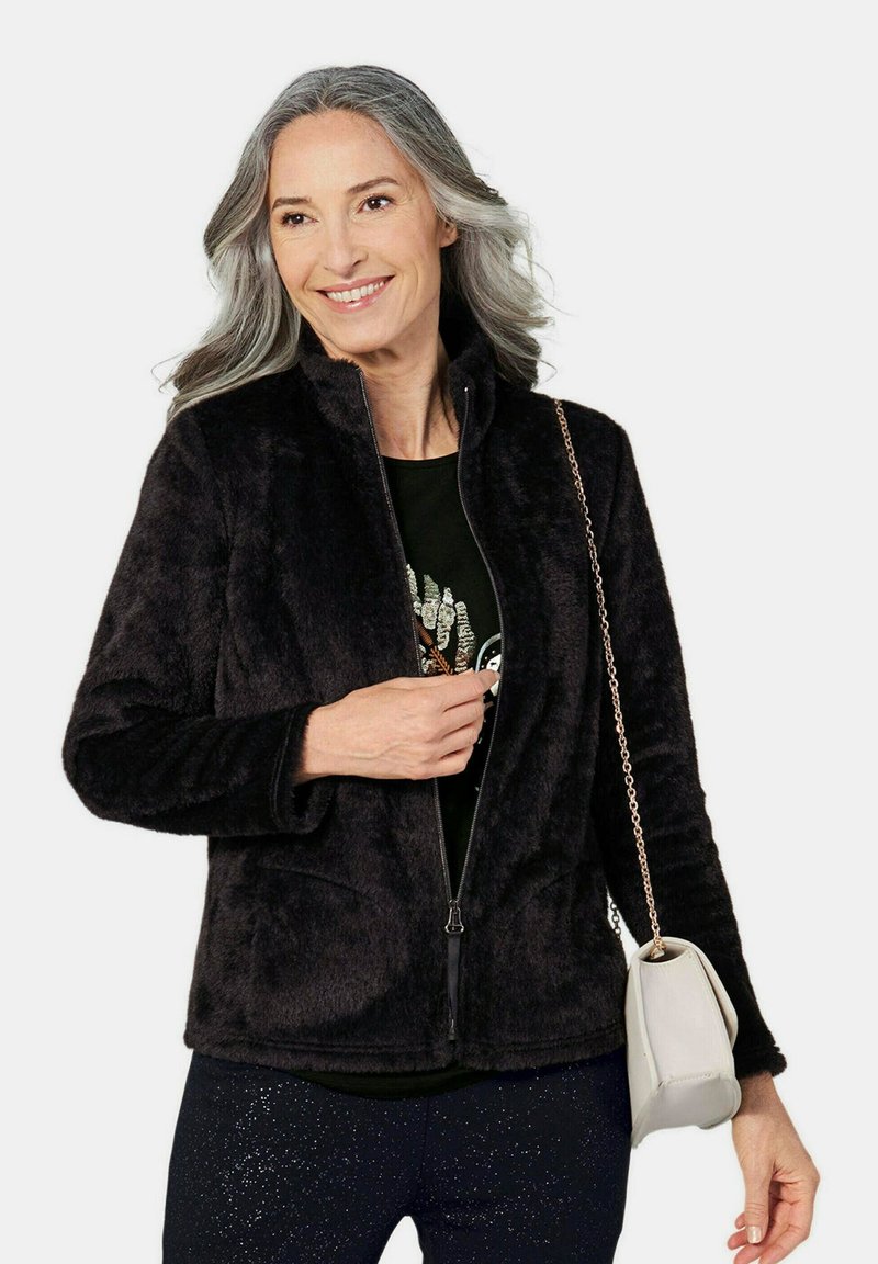 Lachende vrouw met grijs haar draagt een zwart, zacht, ritssluiting jasje, een zwart shirt, glinsterende zwarte broek en draagt een beige tas met een gouden kettingriem.