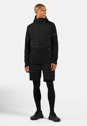 Man draagt een volledig zwart sportief outfit met een jas met capuchon, gewatteerde shorts, zwarte legging en zwarte sneakers, staand en naar de zijkant kijkend.