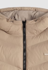 Chaqueta acolchada beige con un alto cuello, cremallera negra y patrones de costura horizontal sutiles. Textura suave y lisa con detalle de etiqueta de logo.