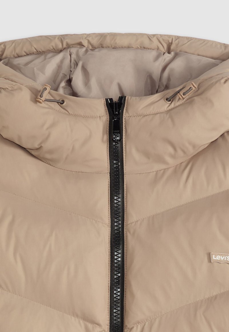 Giacca imbottita beige con colletto alto, cerniera nera e sottili modelli di cucitura orizzontale. Texture morbida e liscia con dettagli del logo.