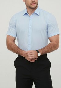 Chemise à manches courtes bleu clair avec un col boutonné et une texture lisse. Associée à un pantalon noir et une montre au poignet gauche.