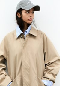 Veste d'extérieur beige avec un col, fermée par un bouton, portée sur une chemise rayée bleue. Une casquette grise complète le look.