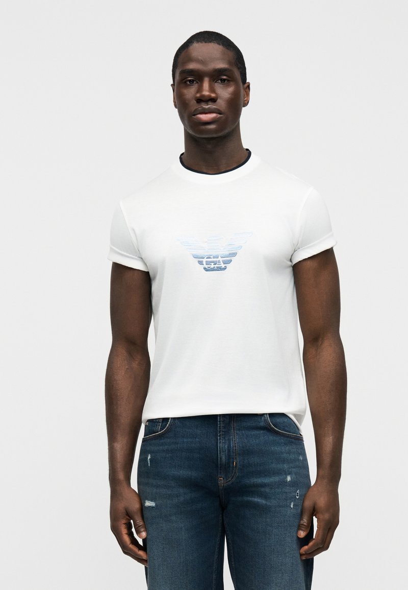 Λευκό κοντομάνικο μπλουζάκι με μπλε λογότυπο με κλίση στο στήθος, σε συνδυασμό με σκούρο μπλε τζιν με φθαρμένες λεπτομέρειες.