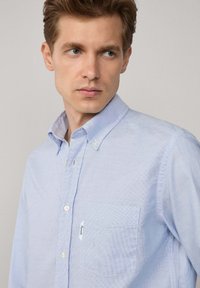 Camisa de botón azul claro con un patrón de cuadros, cuello y un bolsillo en el pecho izquierdo. La tela parece suave y ligeramente texturizada.