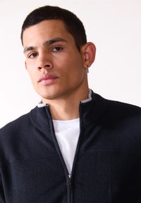 Pulover negru cu fermoar, dintr-un material texturat cu nervuri, cu un guler gri în contrast, purtat peste un tricou alb, având un detaliu elegant al fermoarului.