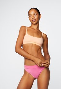 Vrouw die een perzikkleurige bh-top en roze ondergoed draagt, staat tegen een effen lichte achtergrond, kijkt naar de zijkant met handen bij de taille.