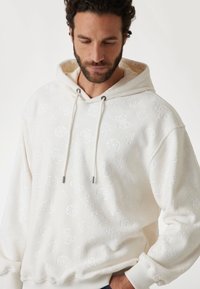 Hvid hættejakke med prægede mønstre, der har snoretræk i hætten, ribbed håndled og en blød, struktureret finish. Accenter i silver-tone hardware.