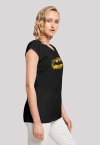 Black cotton t-shirt with short rolled sleeves, featuring a yellow Batman logo centered on the front, worn with white pants.