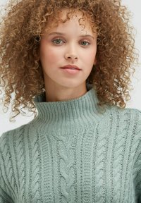 Hellgrüner Pullover mit hohem, geripptem Kragen und Zopfmuster. Strukturierte Oberfläche mit unterschiedlichen Strickdesigns über das gesamte Kleidungsstück.