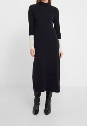 Kobieta ubrana w długą czarną sukienkę z białymi mankietami oraz czarne kozaki za kolano, stojąca na tle jasnego, jednolitego tła.