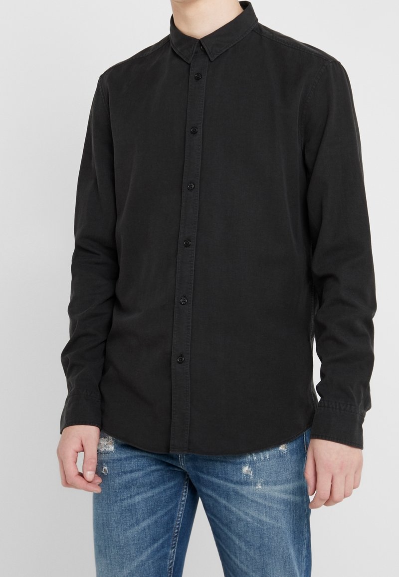 Czarna koszula z długim rękawem z bawełnianej mieszanki, z klasycznym kołnierzykiem, zapięciem na guziki z przodu i mankietami; w połączeniu z jasnoniebieskimi, poszarpanymi dżinsami.