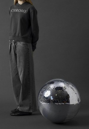 Sweat à capuche noir avec le texte "CHROME", associé à un jean gris ample. Une grosse boule à facettes réfléchissante se tient à proximité sur un fond gris.