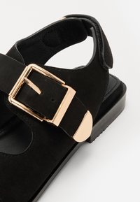 Sorte semskede sandaler med en gullig metallspenne, åpen bak, og en flat, sort såle med rund tå.
