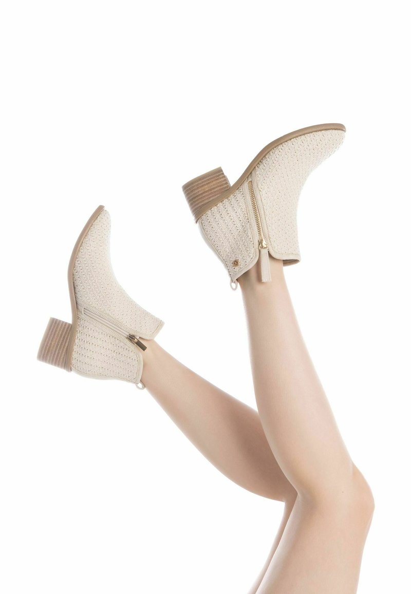 Botas de tobillo beige con parte superior de tela texturizada, cremallera lateral y tacón de bloque de madera. Mostradas con las piernas levantadas sobre un fondo neutro.