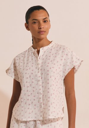 Kobieta ma na sobie białą, krótka, zapinaną na guziki bluzkę z małym różowym kwiatowym wzorem, stojącą na tle jednolitego jasnego tła.