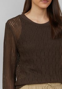 Pull en tricot marron avec un motif texturé et ajouré, doté d'un col rond et de manches longues ; s'accorde avec un pantalon léger à taille cintrée.