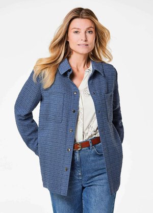 Blauwe, textuurrijke jas met een knoopsluiting aan de voorkant, twee borstzakken en een kraag, gedragen over een bedrukt shirt en blauwe jeans.