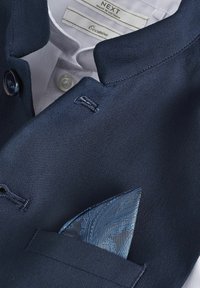 Mornarica blejzer od teksturiranog materijala, s bijelim kragnom košulje. Svijetloplavi paisley maramicom je uredno presavijen u džepu na prsima.