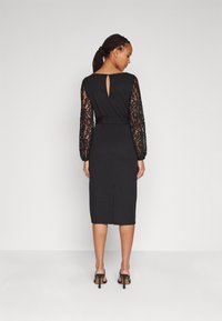 Robe noire avec de longues manches en dentelle, détail en clé au dos, taille ajustée, longueur midi, avec une subtile fente au dos.