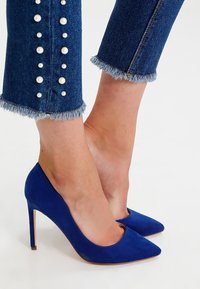 Blå mockaskor med hög klack och spetsig tå, i kombination med denimjeans som har pärldekorationer och fransade fållar.