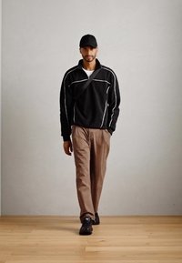 Black zip-up jacket with white accents, beige trousers, and black sneakers. Model wears a black cap, standing on a wooden floor.