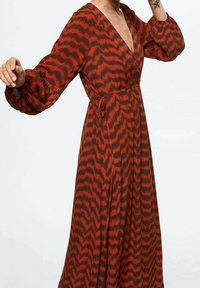 Frau in einem langärmligen, knöchellangen roten Kleid mit schwarzem Zickzackmuster und gebundenem Taillengürtel, die mit einem erhobenen Arm posiert.